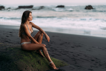 Cute sexy girl in beige swimwear rests on black volcanic sand beach.