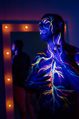 UV patterns body art of the circulatory system on a man's body. On the torso of a muscular athlete, veins and arteries are drawn with fluorescent dyes. Bodybuilder standing by the mirror with lamps.