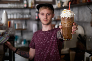 Portrait of a happy young barista man in baseball cap standing behind the bar in trendy city cafe shop table and holding big glass of fresh cold delicious handmade mocaccino, blurred background with