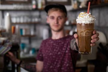 Portrait of a happy young barista man in baseball cap standing behind the bar in trendy city cafe shop table and holding big glass of fresh cold delicious handmade mocaccino, blurred background with