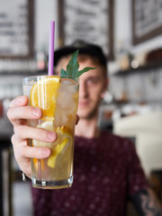 Portrait of a happy young barista man in baseball cap standing behind the bar in trendy city cafe shop table and holding big glass of fresh cold delicious handmade spearmint orange lemonade, blurred