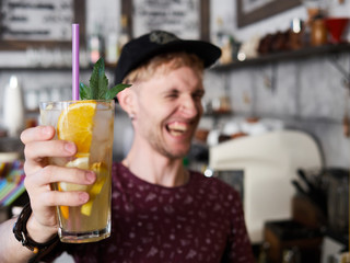 Portrait of a happy young barista man in baseball cap standing behind the bar in trendy city cafe shop table and holding big glass of fresh cold delicious handmade spearmint orange lemonade, blurred
