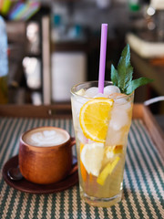 Detail shot of big glass of fresh cold delicious handmade spearmint orange lemonade and brewed cappuccino with latte art in craft clay mug standing on bar tend in cozy city cafe, blurred background