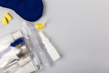Set for flight small bottles with cosmetics, sleep mask, earplugs, nasal spray on gray background. Top view, copy space