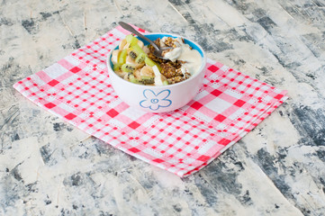 Obraz na płótnie Canvas Granola with Greek yogurt kiwi and banana in bowl on a pink napkin, view from the side. Piece of dessert on a spoon. Fitness diet for weight loss, proper and tasty food