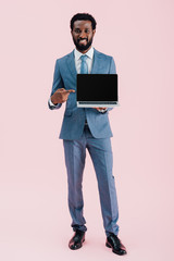 smiling african american businessman pointing at laptop with blank screen isolated on pink