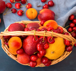 Wicker basket with seasonal summer fruits and berries. Ripe juicy nectarines apricots sweet cherries scattered on blue cotton towel. Vitamins healthy balanced diet concept