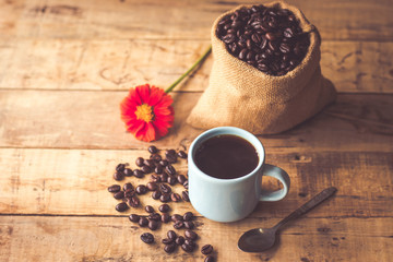 Black coffee In the cup sky blue color have roasted coffee beans are In a cloth bag sack and on table with, still have red flowers and stainless steel spoon all placed on a wooden table.