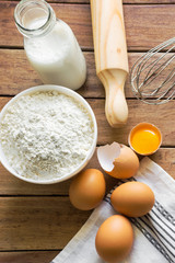 Basic baking cooking ingredients organic free ranch brown eggs with cracked shell bright yellow yolk milk in reusable glass bottle flour rolling pin whisk cotton towel on wooden kitchen table