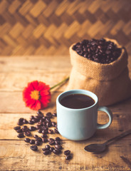 Black coffee In the cup sky blue color have roasted coffee beans are In a cloth bag sack and on table with, still have red flowers and stainless steel spoon all placed on a wooden table.