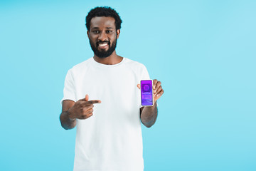 smiling african american man pointing at smartphone with shopping app, isolated on blue