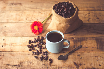 Black coffee In the cup sky blue color have roasted coffee beans are In a cloth bag sack and on table with, still have red flowers and stainless steel spoon all placed on a wooden table.