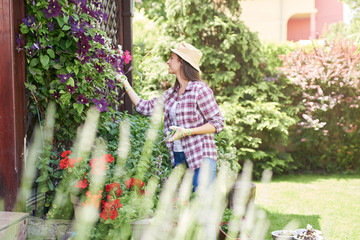 Beautiful smiling Caucasian brunette with hat, gardening gloves and in working clothes taking care of her flowers in backyard.