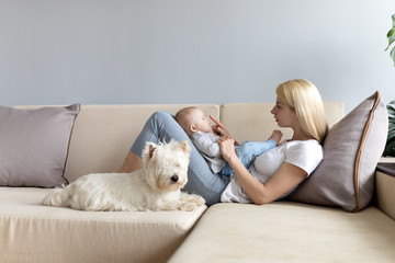 Young mother and child in her hands happy with white westie west highland white terrier dog on a white sofa with grey wallss miling and playng