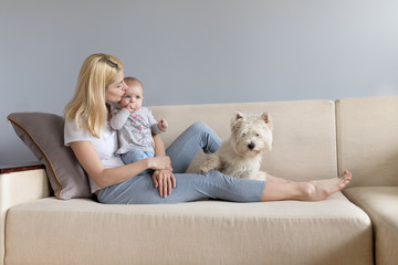 Young mother and child in her hands happy with white westie west highland white terrier dog on a white sofa with grey wallss miling and playng