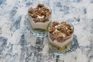 Granola, kiwi, banana and greek yogurt in glass cups on gray concrete