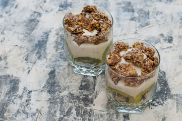Granola, kiwi, banana and greek yogurt in glass cups on gray concrete