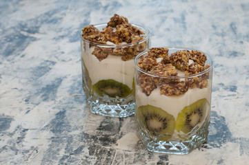 Granola, kiwi, banana and greek yogurt in two glass cups on gray concrete