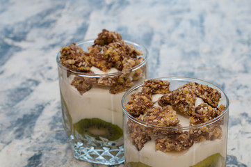 Granola, kiwi, banana and greek yogurt in two glass cups on gray concrete