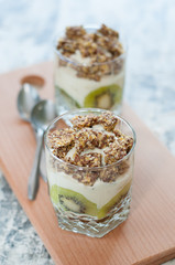 Granola, kiwi, banana and Greek yogurt in two glass cups on a wooden board gray concrete, with spoons, copy space, vertical format. Fitness, figure, body and healthy food