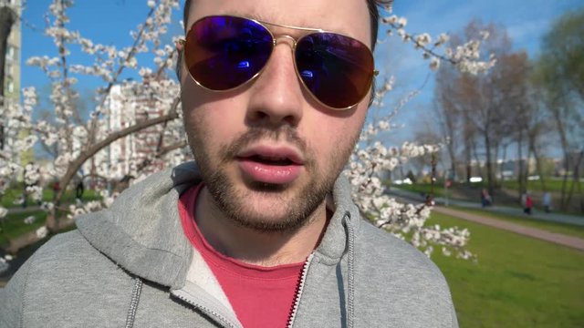 Aggressive man in sunglasses looks at the camera and speaks. Adult man stands near the apricot tree in city park and talks into the camera