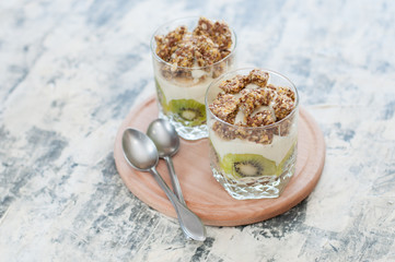 Obraz na płótnie Canvas Dessert granola, Greek yogurt, kiwi and banana in two glass cups with spoons on wooden round board, gray concrete, close-up. Fitness, figure, body and healthy food