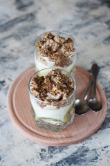 Dessert granola, greek yogurt, kiwi and banana in two glass cups with spoons on a wooden round board, gray concrete, close-up vertical frame format. Fitness, figure, body and healthy food