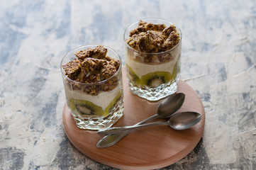 Dessert granola, Greek yogurt, kiwi and banana in two glass cups with spoons on a wooden round board, gray concrete, close-up. Fitness, figure, body and healthy food