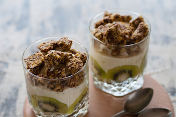 Dessert granola, Greek yogurt, kiwi and banana in two glass cups with spoons on a wooden round board, gray concrete, close-up. Fitness, figure, body and healthy food