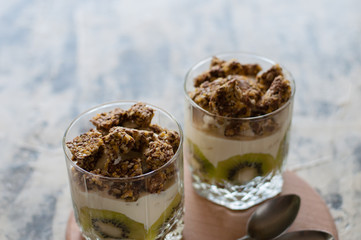 Dessert granola, Greek yogurt, kiwi and banana in two glass cups with spoons on a wooden round board, gray concrete, close-up. Fitness, figure, body and healthy food