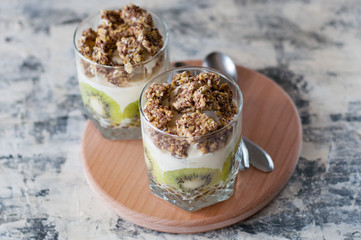 Dessert granola, Greek yogurt, kiwi and banana in two glass cups with spoons on wooden round board, gray concrete, copy space, horizontal format. Fitness, figure, body and healthy food
