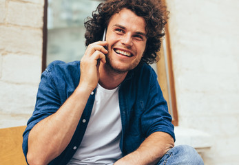 Portrait of happy young man stitting on the bench outdoors and talking on mobile phone. Happy male with curly hair resting outside making a call on his cell phone in the city street. Lifestyle, people