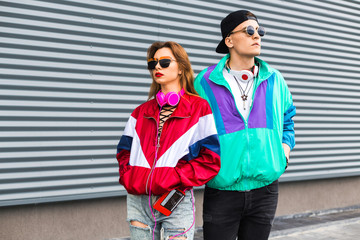 Back in time 90s 80s. Stylish young man in a retro jacket and a girl in red and with a vintage cassette player, against a steel wall, fashion trends, a street image