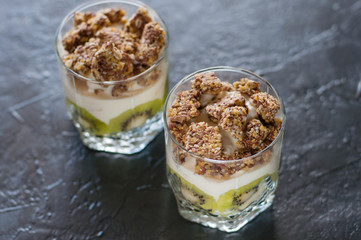 Greek yogurt, granola, banana and kiwi. Two desserts in glass cups on dark background. Concept body and healthy food