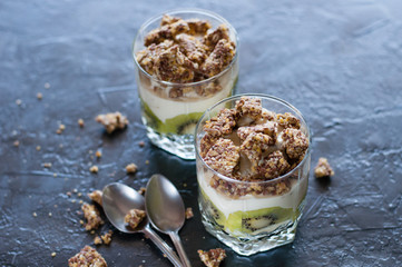 Greek yogurt, granola, banana and kiwi. Desserts in glass with spoons on a dark background. Concept body and healthy food