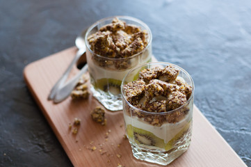 Greek yogurt, granola, banana and kiwi. Desserts in glass cups with spoons on wooden platform, dark background, view from the top. Concept body and healthy food