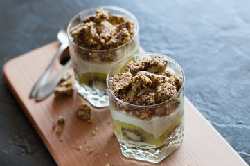 Greek yogurt, granola, banana and kiwi. Desserts in glass cups with spoons on wooden platform, dark background, view from the top. Concept body and healthy food
