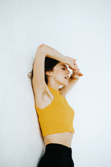 portrait of brunette woman wearing a yellow top leaning against the wall with arms raised and closed eyes