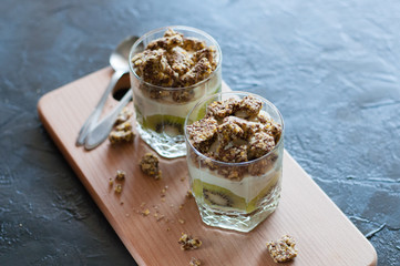 Greek yogurt, granola, banana and kiwi. Desserts in glass cups with spoons on wooden platform, dark background, view from the top. Concept body and healthy food