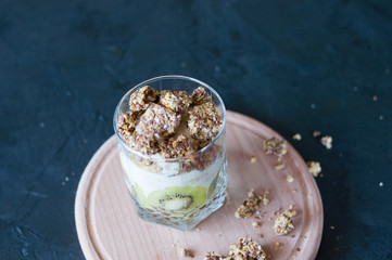 Granola, banana, kiwi and greek yogurt. Dessert in glass cup on a wooden round board, dark background. Concept body and healthy food