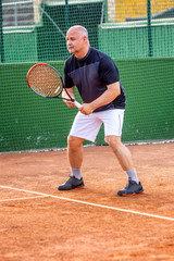 A middle-aged bald man plays tennis on the outdoor court. Sunny day. Vertical.