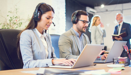 Young business woman with headset using laptop at her workplace in office. Female sales agent talking with client using headphones and microphone and working on laptop at her workplace..