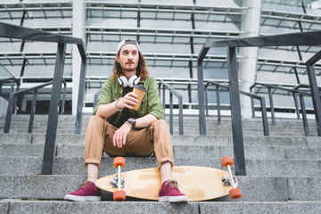 Low angle view of calm man with paper cup in hand looking away, sitting on stairs near skateboard