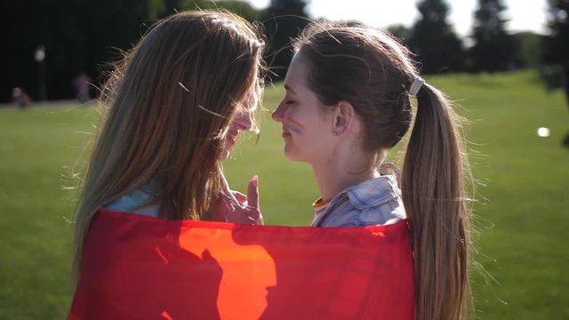 Close-up portrait of gay couple standing cuddling and touching foreheads wrapped in rainbow flag in countryside park. Smiling females with lgbt flag painted on cheeks expressing tenderness and love