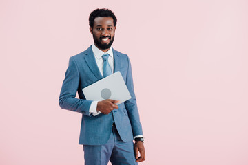 cheerful african american businessman holding laptop isolated on pink