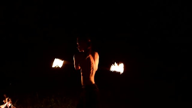 Low key. Young male with long hair and bare torso rotates burning torch outdoors on a black night video slow motion. Modern fakir does tricks with a burning staff