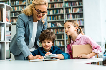 cheerful teacher in glasses standing near cute kids reading book in library
