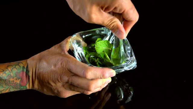 This close-up panning video of a tattooed asian males hands muddling mint leaves in a beautiful clear crystal mortar and pestle on a clack background.