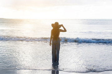 Young woman in hat standing in water on sandy beach and looking to a sky.