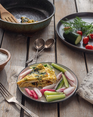 Breakfast of scrambled eggs with spinach and cutting of vegetables, whole vegetables and a pan with omelette remains on a rustic table board